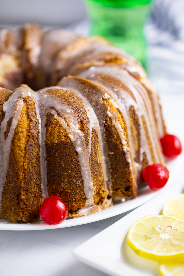 Close up shot of 7Up pound cake drizzled with glaze with maraschino cherries on a white platter, next to lemon slices on a rectangle plate and a bottle of 7Up in the background