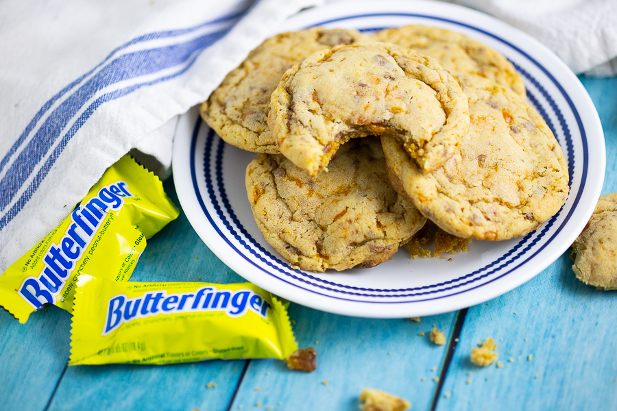 Butterfinger cookies stacked on a white plate next to mini Butterfingers and a white and blue linen on an aqua wood background