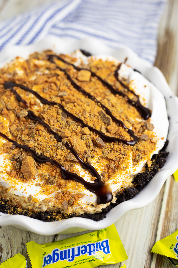 A whole Butterfinger pie in a white ruffled pie dish with Butterfingers and a white and blue linen around it.