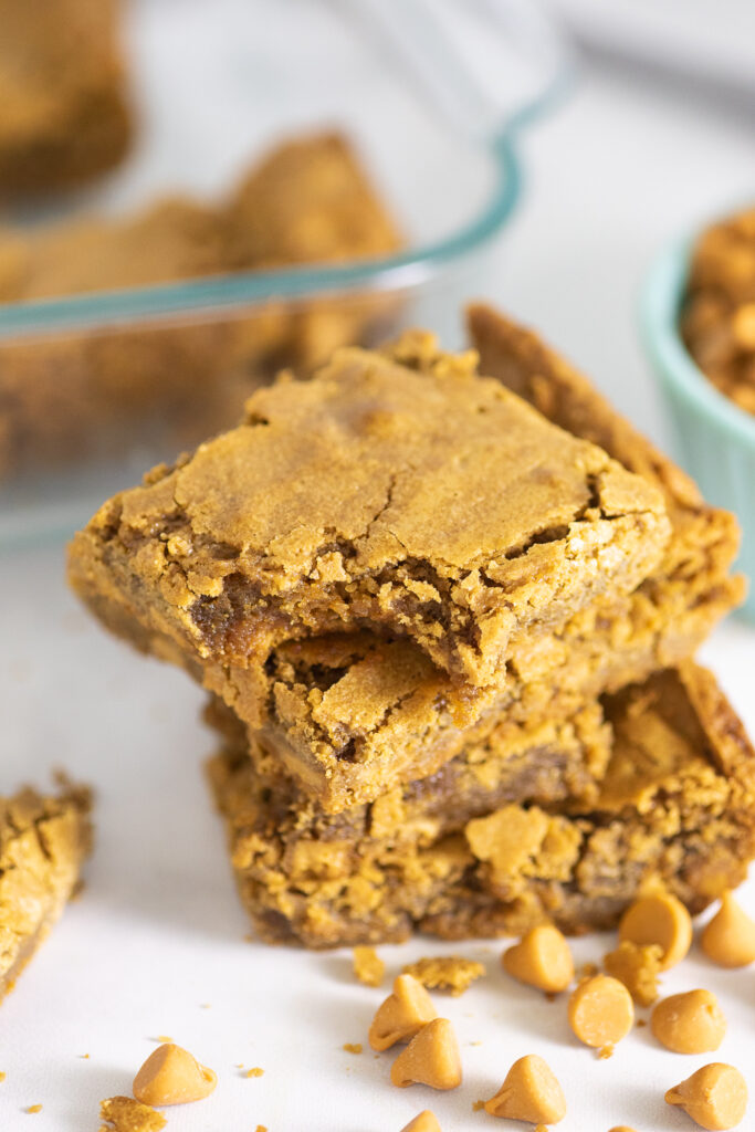 Stack of 4 butterscotch blondies with a bite taken out of the top one on a marble counter with a glass baking dish and small bowl of butterscotch chips behind it