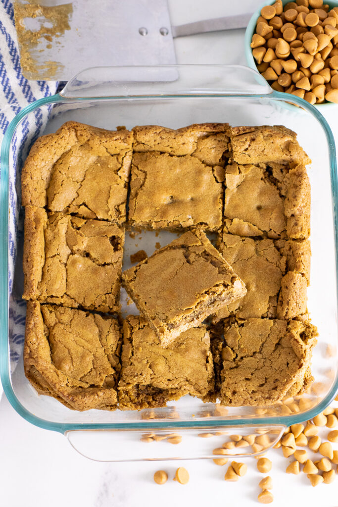 Glass dish with butterscotch blondies cut into 9 bars. Dish is on a marble counter surrounded by a metal spatula, a small cup of butterscotch chips, and loose butterscotch chips scattered around.