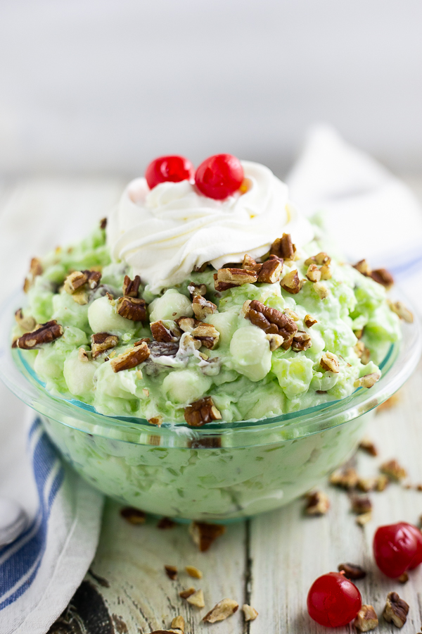 Glass bowl filled with Watergate Salad topped with whipped cream and cherries on a rustic white background.