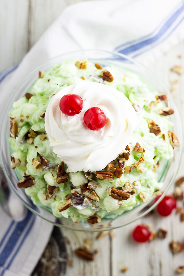 Overhead view of Watergate salad topped with chopped pecans, whipped cream, and maraschino cherries in a glass bowl on a white rustic wooden background.