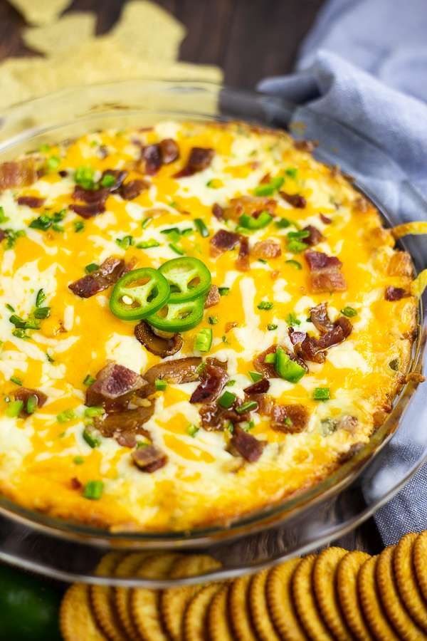 Jalapeno popper dip in a glass pie dish surrounded by chips, crackers, and a light blue linen