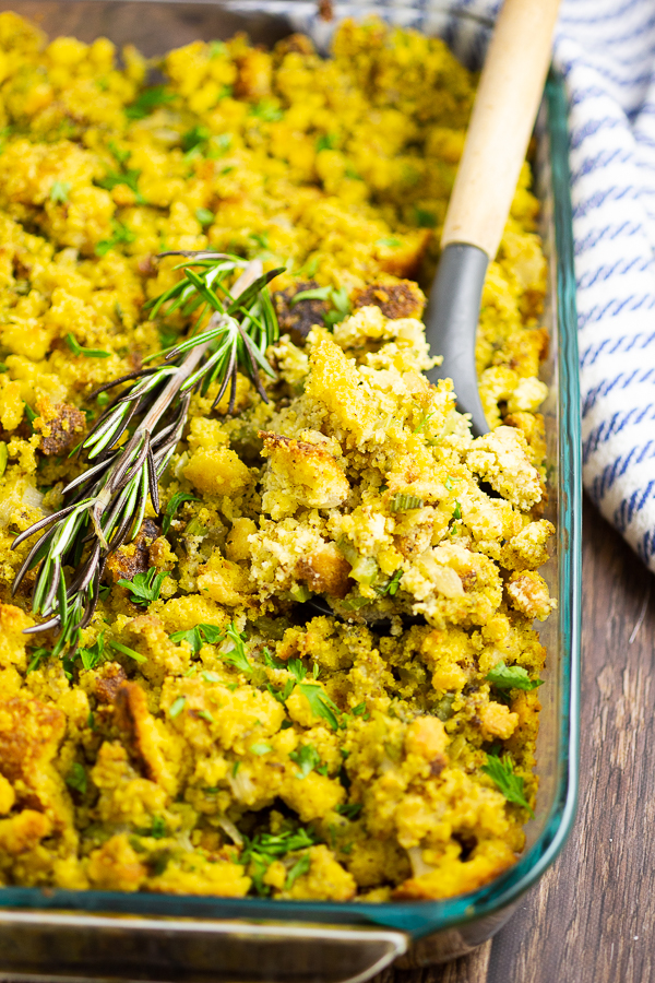 Cornbread dressing in a glass dish with a sprig of rosemary and a wooden spoon in the middle.