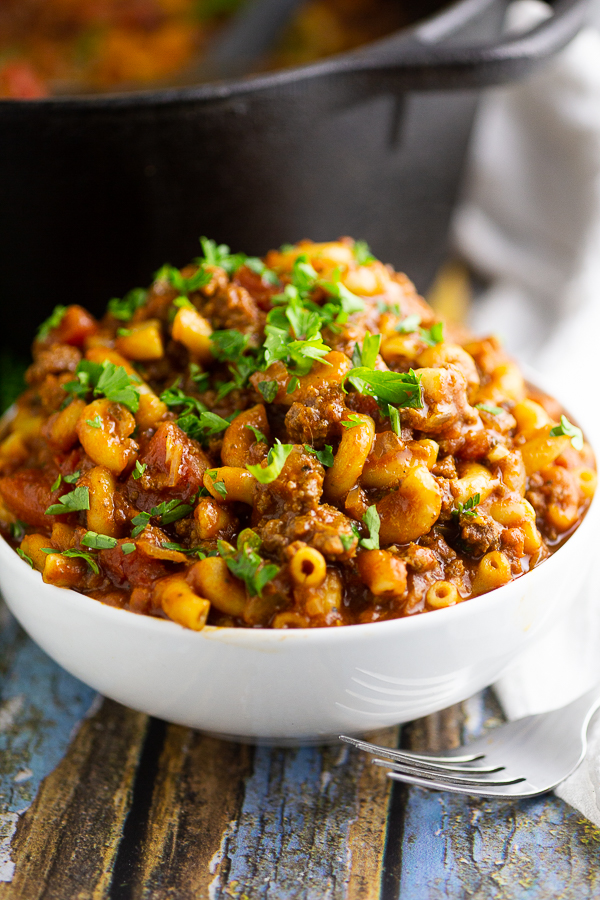 American goulash in a white bowl topped with fresh parsley sitting in front of a cast iron dutch oven