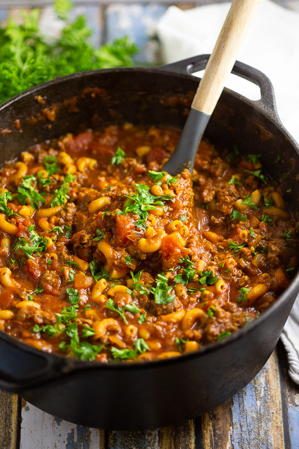 American goulash in a cast iron dutch oven with a ladle in it topped with fresh parsley.