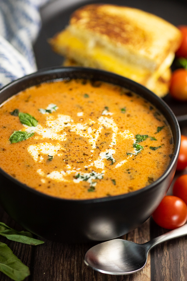 Black bowl full of creamy tomato basil soup topped with fresh chopped basil and cream swirls with a spoon in front, cherry tomatoes next to it, and a grilled cheese sandwich behind