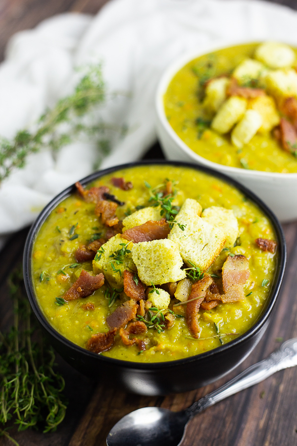 Split pea soup in a black matte bowl topped with bacon, croutons, and thyme leaves with a second bowl behind.