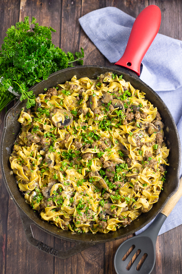 Ground beef stroganoff in a cast iron skillet next to a wooden spoon, fresh parsley, and a canvas linen on a rustic wood background.