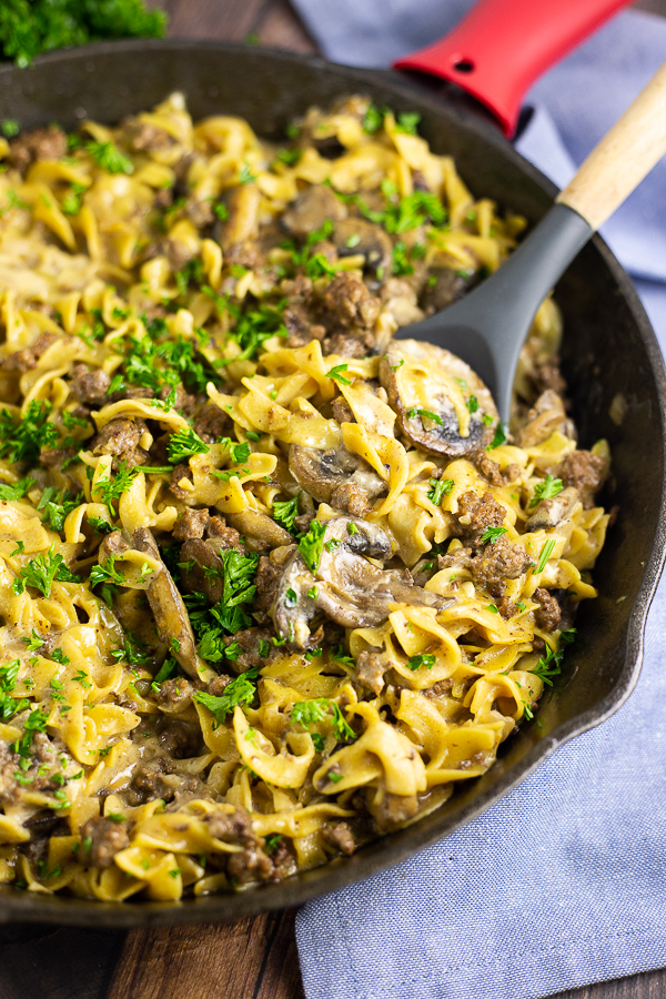 Ground beef stroganoff in a cast iron skillet with a spoon in the middle, a blue canvas linen in the background.