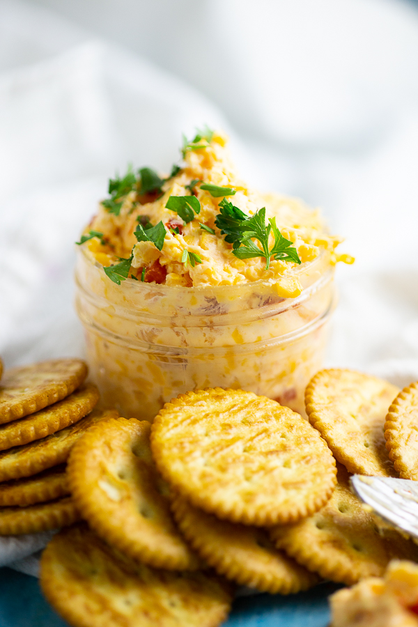 Homemade pimento cheese in a small mason jar, garnished with fresh parsley. The jar is surrounded by butter crackers.