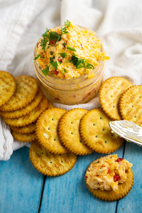 Overhead picture of pimento cheese in a small jar garnished with parsley, surrounded by butter crackers. One cracker has pimento cheese spread on it.