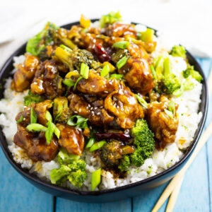 General Tso's Chicken in a bowl on top of white rice with steamed broccoli topped with sliced green onions. The bowl is on a bright blue background next to chopsticks.