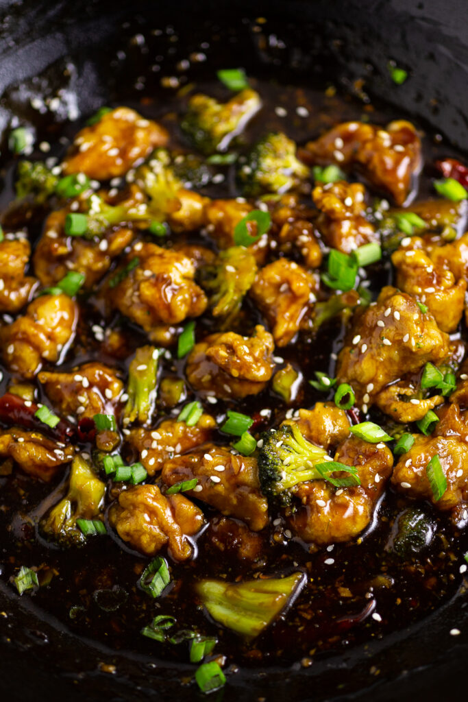 Close up of General Tso's Chicken with broccoli in a large cast iron wok topped with sliced green onions.