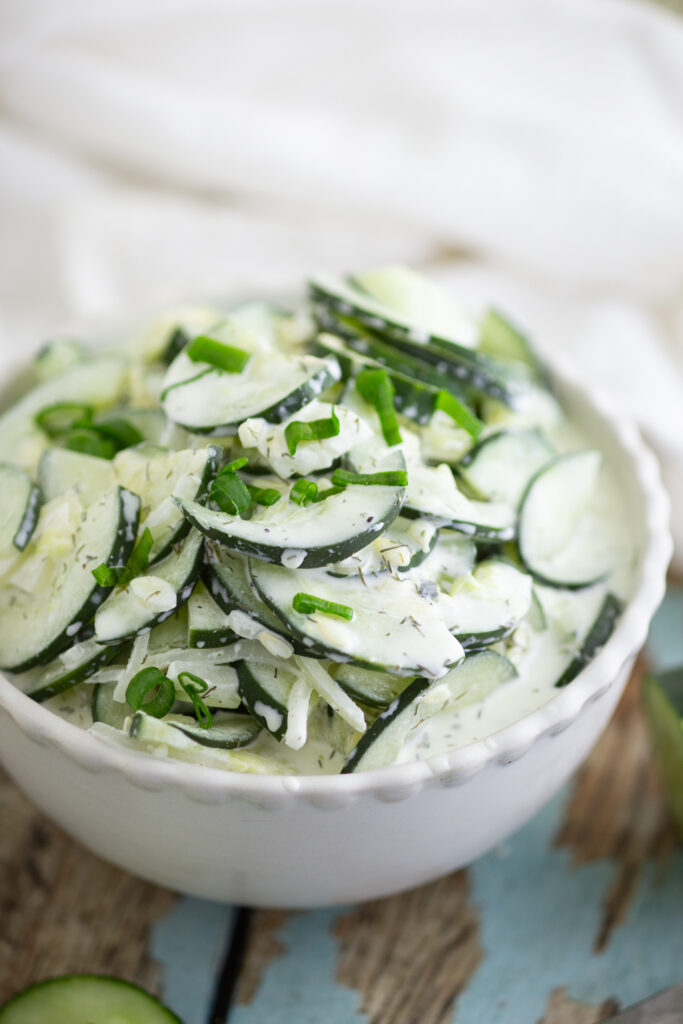 Creamy Cucumber salad topped with fresh chives in a white ceramic bowl sitting on a rustic wood background with a linen napkin behind the bowl.