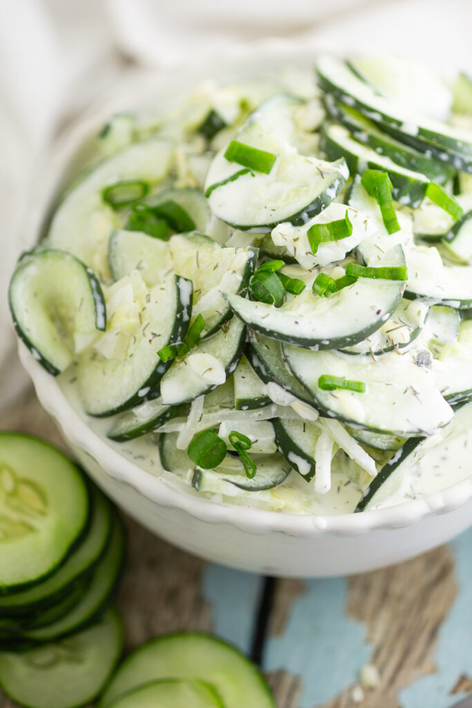 Close up of creamy cucumber salad topped with freshly chopped chives in a white ceramic bowl next to sliced cucumbers on a rustic wood background.