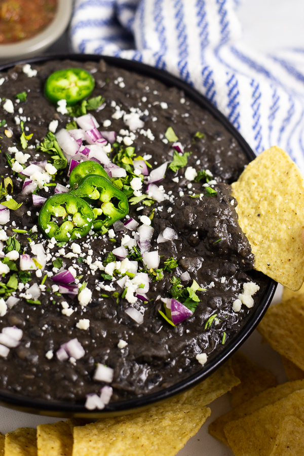 Refried black beans topped with cotija cheese, jalapenos, and red onion in a black matte bowl with a chip dipping in