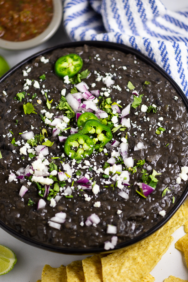 Close up of refried black beans in a black bowl surrounded by a striped linen, tortilla chips, a cup of salsa, and a lime.