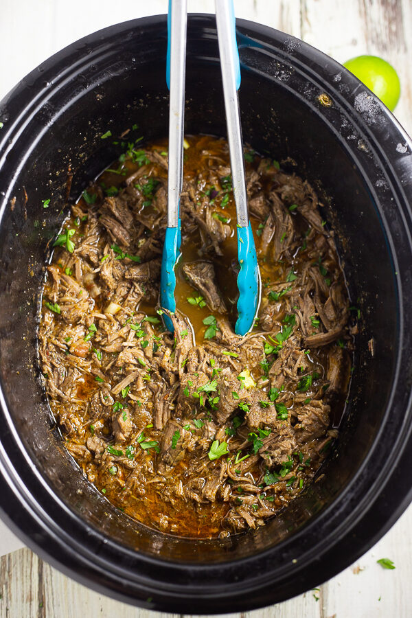Shredded Chipotle barbacoa in a Crock pot with tongs in the middle garnished with fresh cilantro