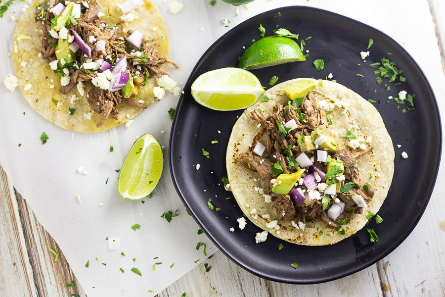 Open barbacoa street taco on a black matte plate with fresh lime wedges and another taco in the background.