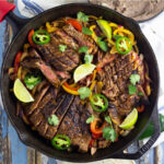 Overhead view of fajita seasoned skirt steak on a bed of sauteed bell peppers and onions topped with fresh jalapeno slices and lime wedges