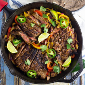Overhead view of fajita seasoned skirt steak on a bed of sauteed bell peppers and onions topped with fresh jalapeno slices and lime wedges