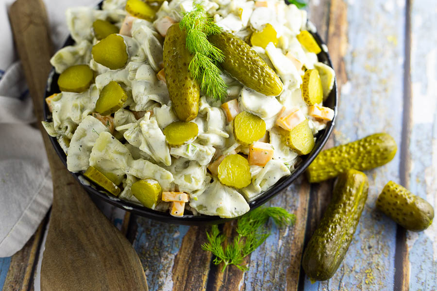 Horizontal image of dill pickle pasta salad in a black bowl topped with 2 baby dills and a sprig of fresh dill. Bowl is placed on a rustic wood background with fresh herbs, 2 more baby dill pickles, and a wooden spoon