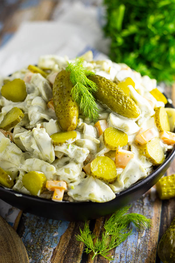 Dill pickle pasta salad in a black bowl topped with 2 baby dills and a sprig of fresh dill. Bowl is placed on a rustic wood background with fresh herbs and a wooden spoon