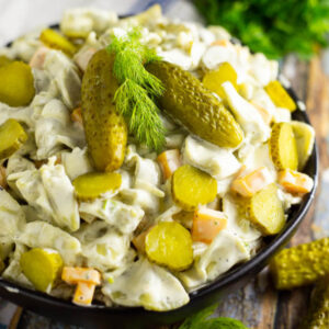 Square image of dill pickle pasta salad in a black bowl topped with 2 baby dills and a sprig of fresh dill. Bowl is placed on a rustic wood background with fresh herbs and a wooden spoon