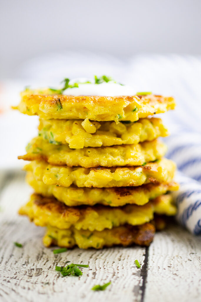 Stack of corn fritters topped with sour cream and freshly chopped chives on a rustic wood background.