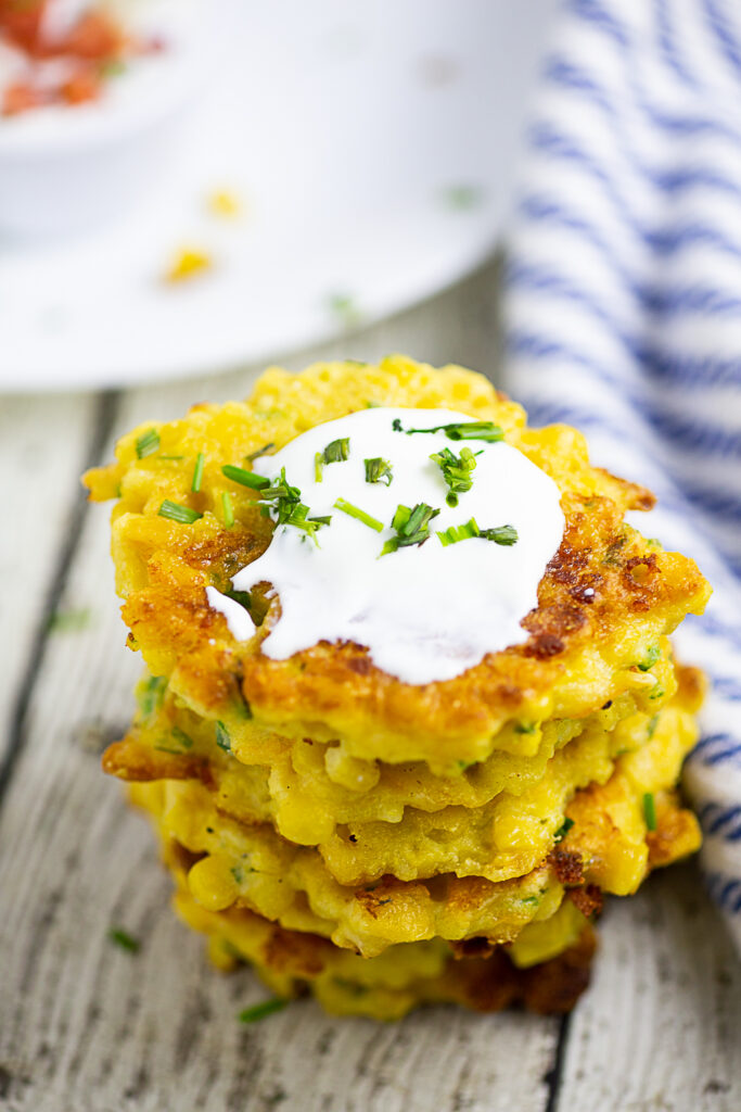 An overhead view of a stack of corn fritters on a rustic wood backdrop. The top fritter is topped with sour cream and freshly chopped chives.