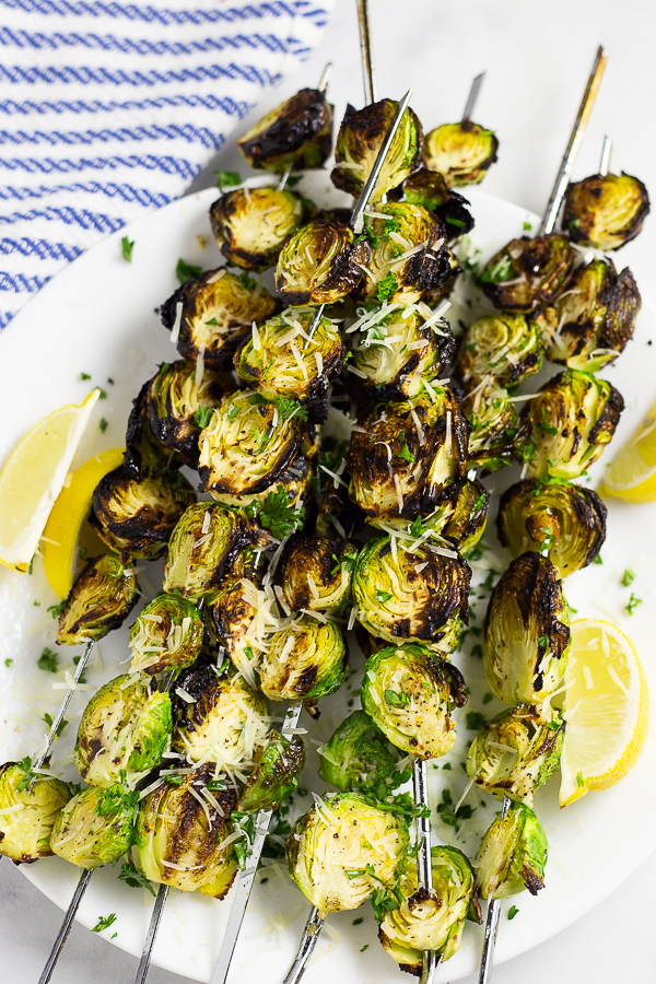 Grilled brussels sprouts on skewers on a white plate with lemon wedges and a striped linen in the background