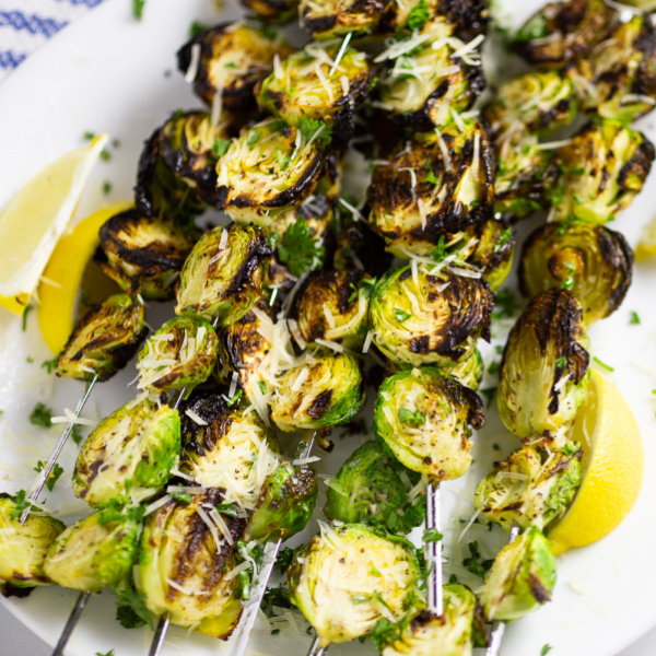 Grilled brussels sprouts on skewers on a plate with lemon wedges topped with grated Parmesan