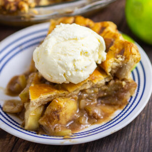 Slice of apple pie on a small plate topped with a scoop of vanilla ice cream. Green apple and pie in a pie dish are behind.