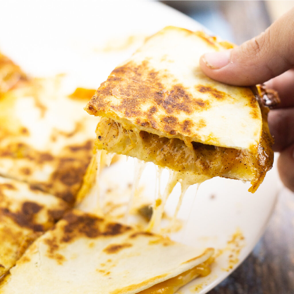 Hand picking up a quarter of a gooey, cheesy quesadilla from a white plate.