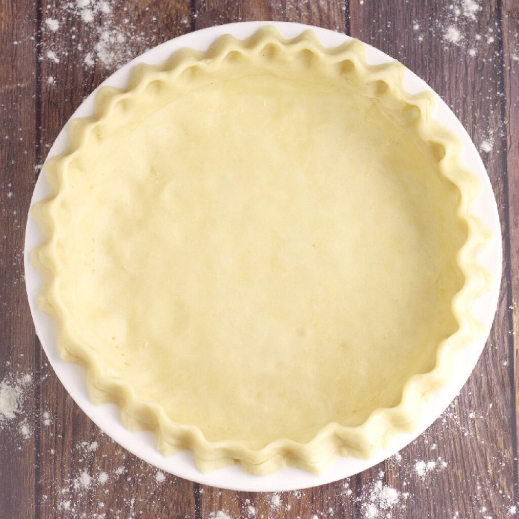 Unbaked pie crust fluted in a white dish on a rustic wood background with flour on it