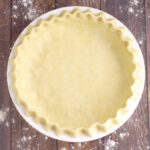 Unbaked pie crust fluted in a white dish on a rustic wood background with flour on it