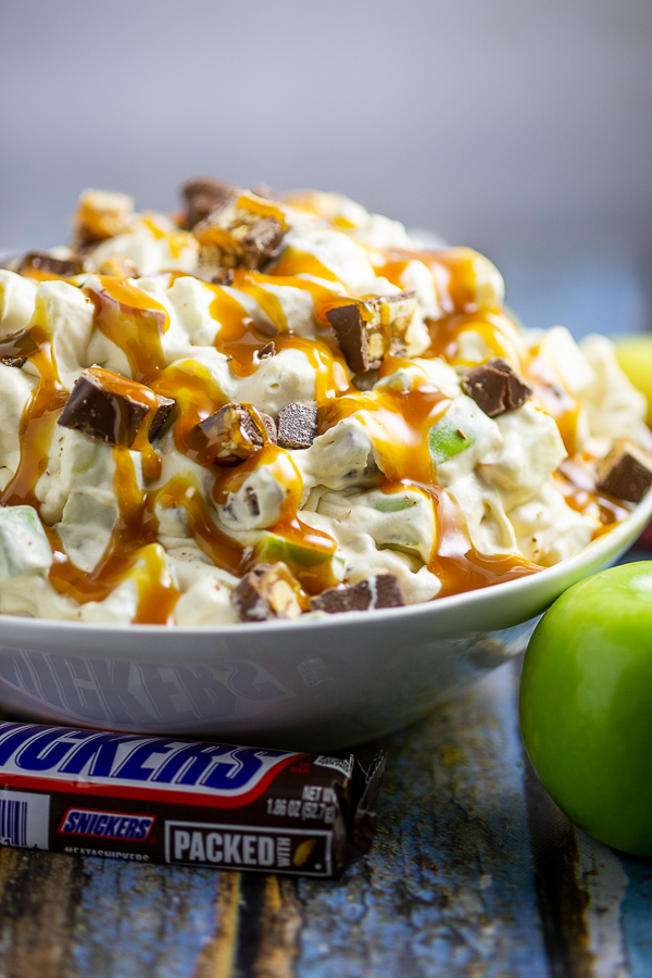 Snickers Caramel Apple Salad drizzled with caramel sauce in a large bowl next to a wrapped Snickers bar and Granny Smith Apple on a rustic wood backdrop