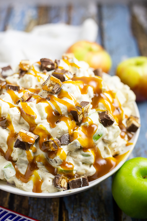 Overhead photo of half a bowl of Snickers Caramel Apple Salad drizzled in caramel sauce next to a Granny Smith apple and 2 Honeycrisp apples