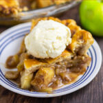 Slice of apple pie on a small plate topped with a scoop of vanilla ice cream. Green apple and pie in a pie dish are behind.