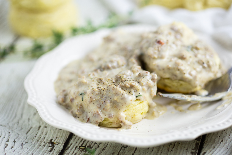 Side view of biscuits on a white plate with Southern sausage gravy dripping down, a fork on the plate, and more biscuits and thyme behind them