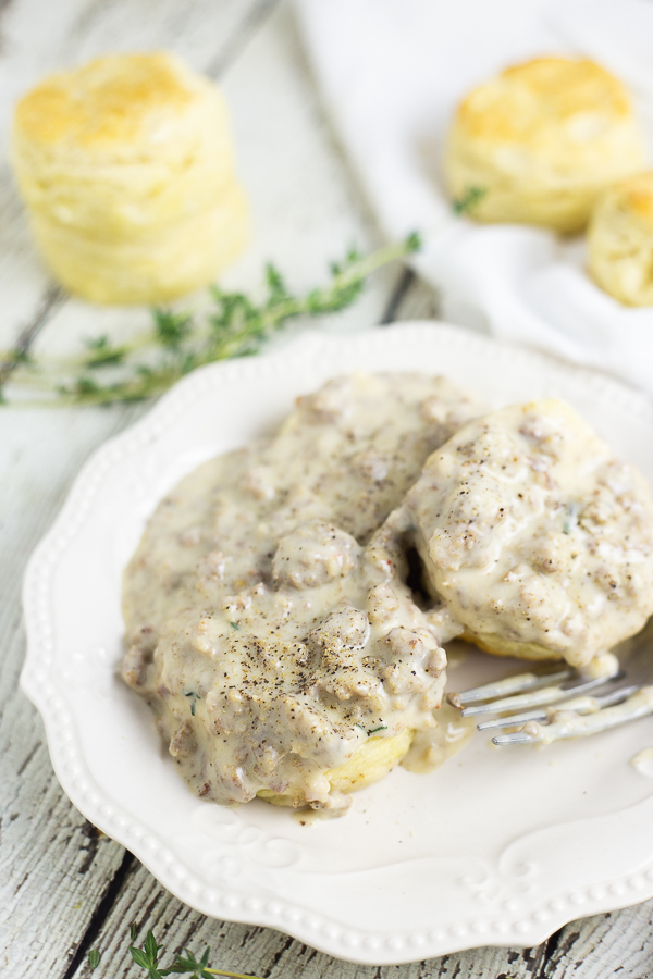 Sausage gravy on buttermilk biscuits on a small plate with more biscuits and thyme in the background