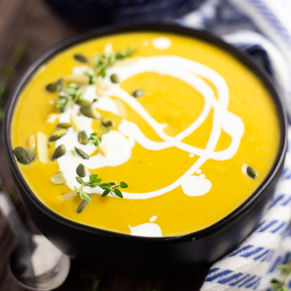 Butternut squash bisque topped with a drizzle of cream with some pepitas and a sprig of thyme. The soup is in a matte black bowl sitting on a rustic wood background with a striped linen