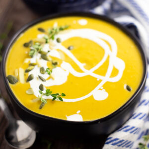 Butternut squash bisque topped with a drizzle of cream with some pepitas and a sprig of thyme. The soup is in a matte black bowl sitting on a rustic wood background with a striped linen