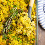 Cornbread dressing in a glass dish with a sprig of rosemary and a wooden spoon in the middle.