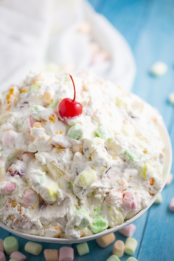 Angle picture of a big bowl of Ambrosia Salad with a cherry on top with colorful mini marshmallows scattered around and a white linen in the background.