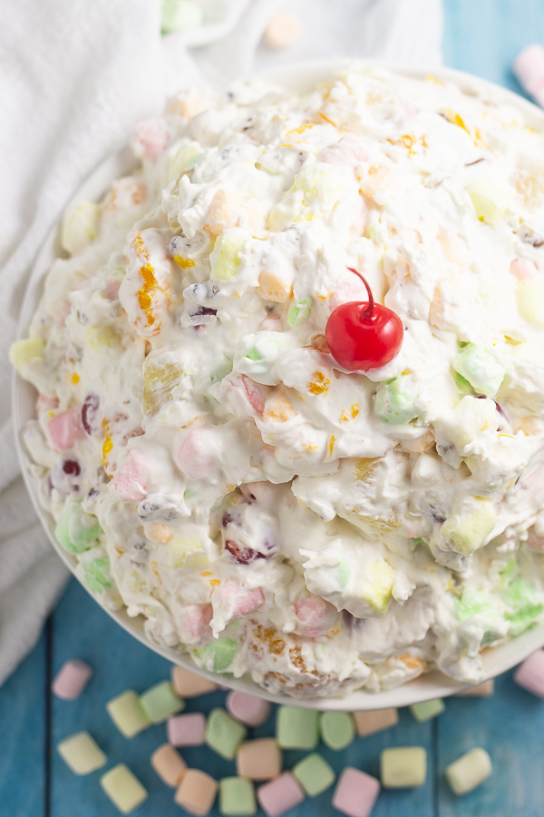 Overhead picture of a big bowl of ambrosia salad with a maraschino cherry on top. The bowl is surrounded by mini marshmallows and next to a white linen