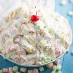 A big bowl of Ambrosia salad with a cherry on top with mini marshmallows scattered around it.