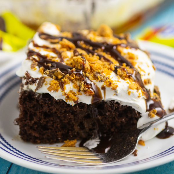 A slice of Butterfinger cake on a small blue striped place with large cake in the background with Butterfinger candy bars